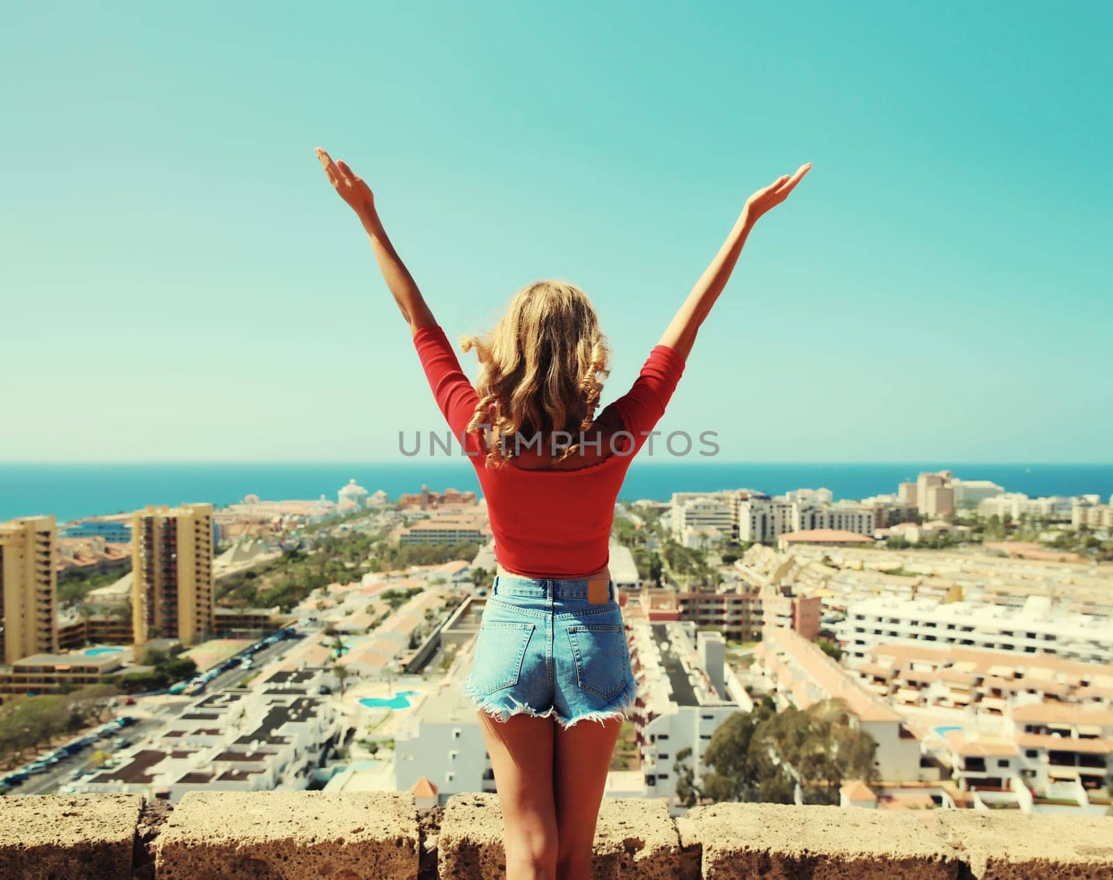 Summer vacation, beautiful happy young woman tourist raising her hands up on top of the mountain looking around the city in Tenerife, Canary Islands, Spain. Aerial top view.