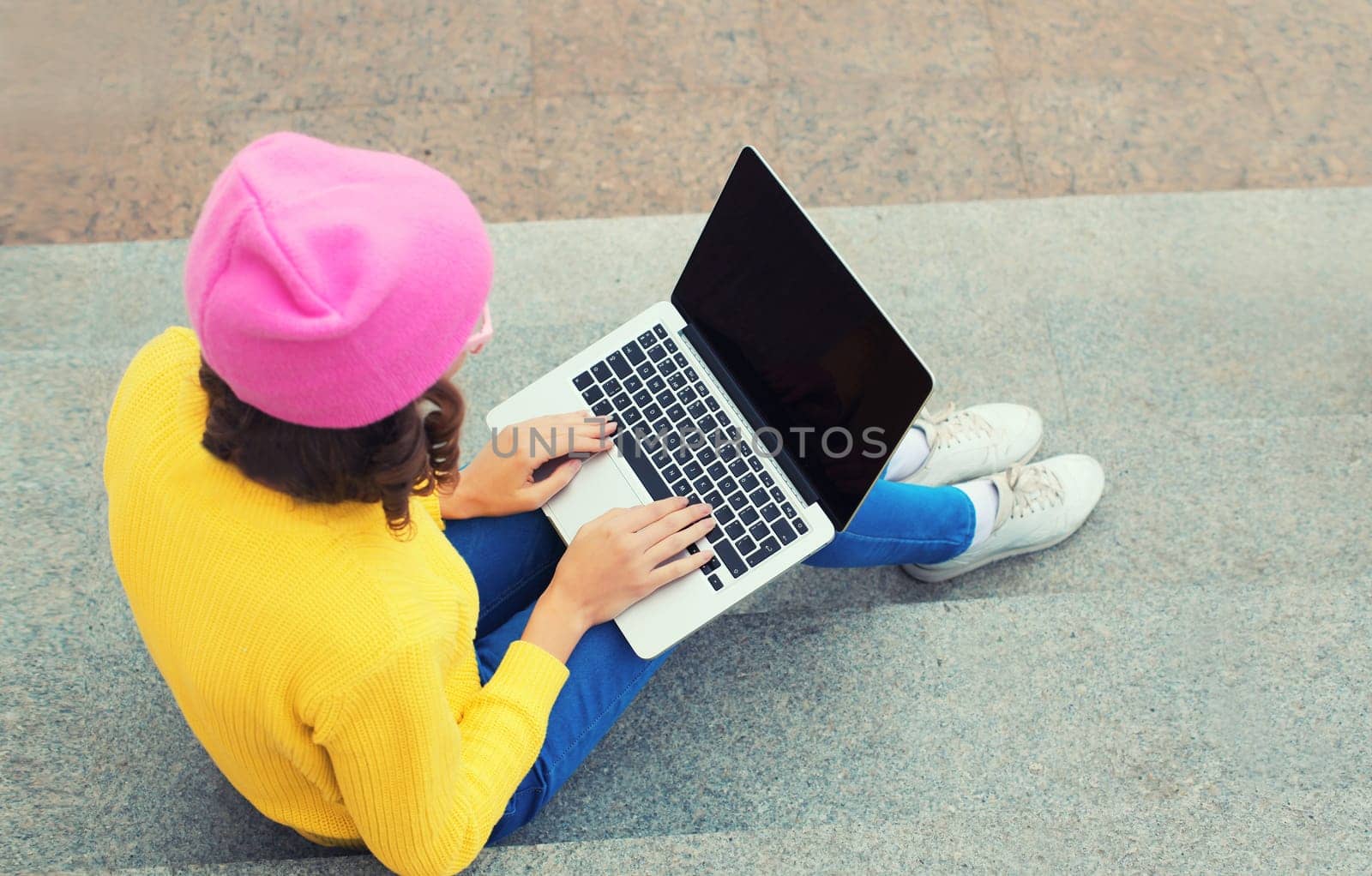 Stylish modern young woman working with laptop in colorful clothes in the city