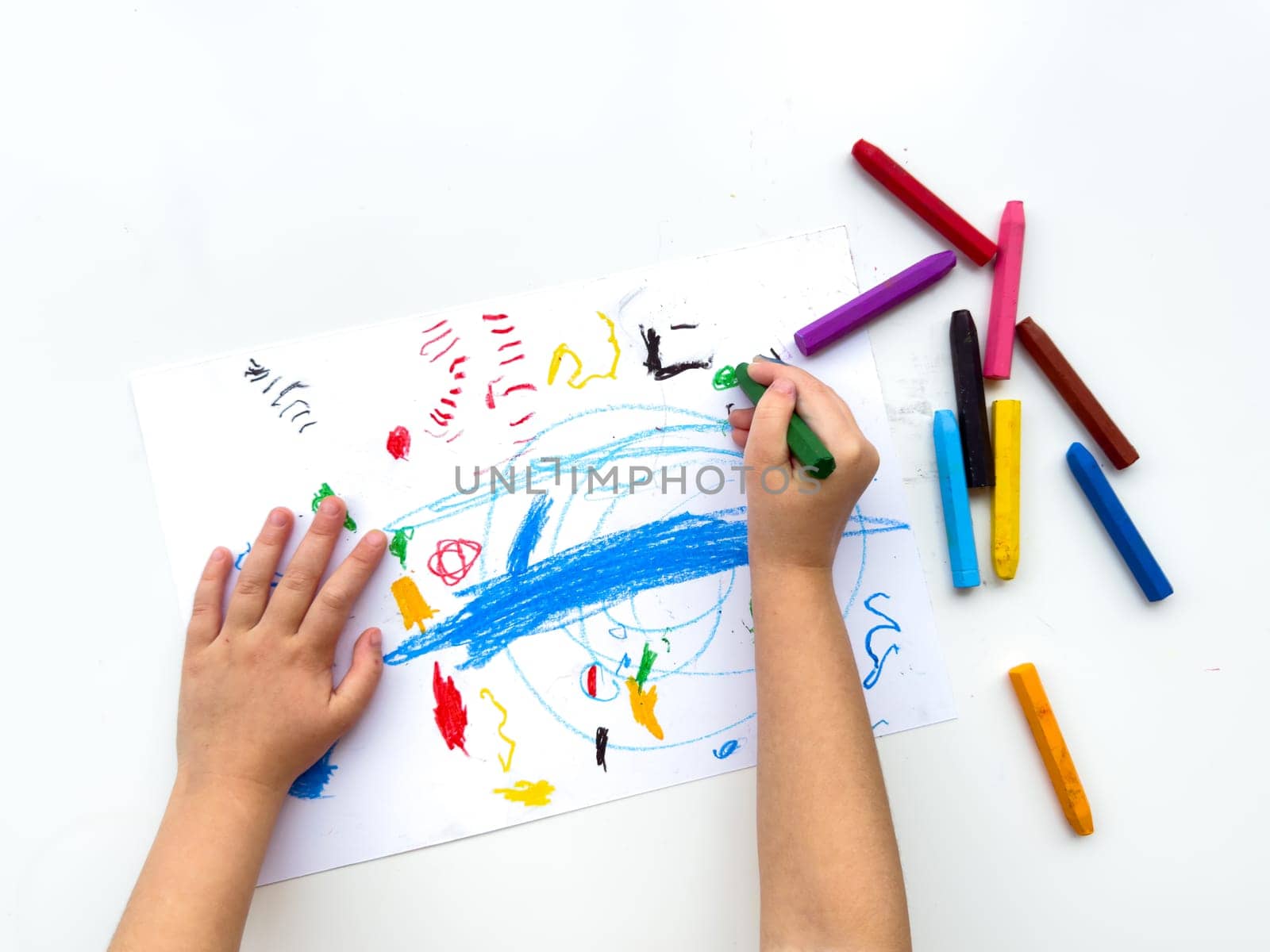 Childs hands drawing with colorful wax crayons on white paper, top view. Creative art concept for educational and developmental activities. High quality photo