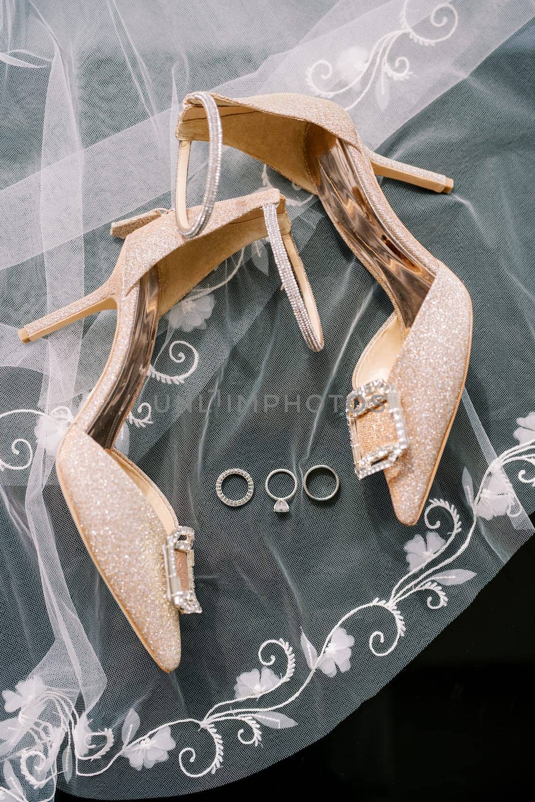 Wedding rings lie on an embroidered veil near the bride golden high-heeled shoes by Nadtochiy