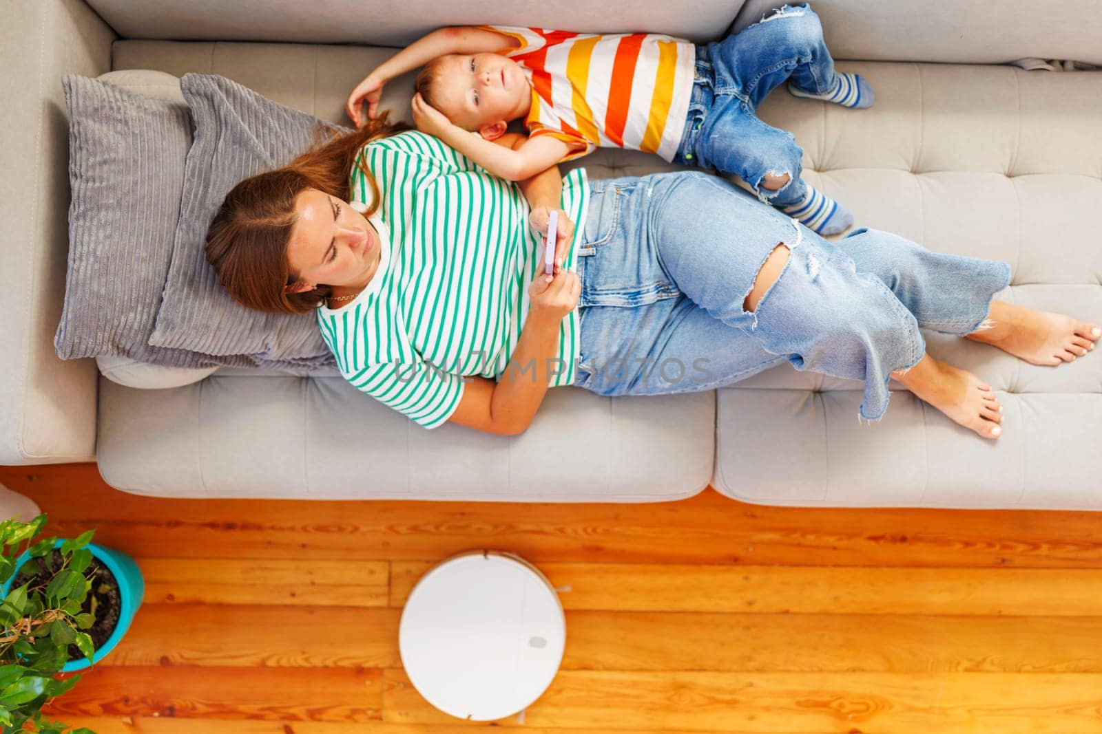 Mother and Child Relaxing on Couch with Smartphone and Robot Vacuum by andreyz