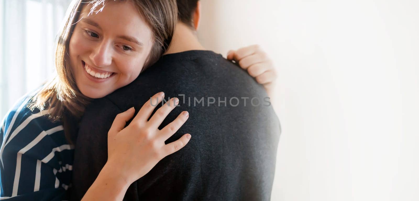 Happy young girl warmly hugs her boyfriend, rejoices at the meeting, stands near the window, smiley face in close-up, woman starts a romantic relationship. Husband and wife feel united, in a harmony.