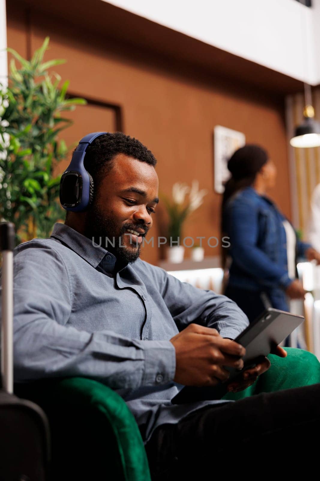 Smiling African American man businessman in headphones using digital tablet checking email during business trip, black guy hotel guest browsing internet while waiting for check-in in lobby