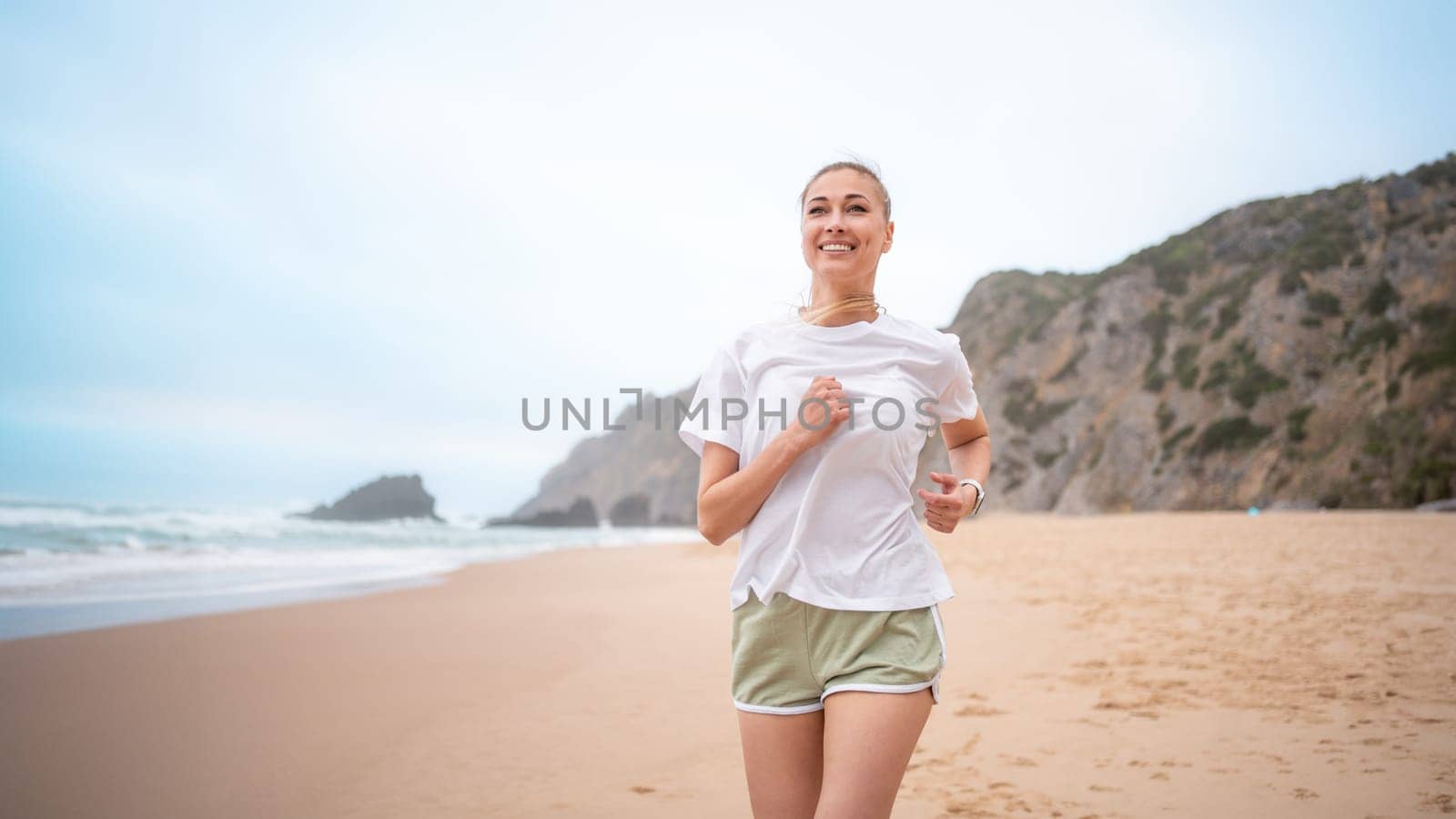 Smiling young woman jogging on sandy beach along sea waves by andreonegin