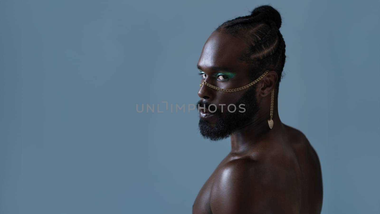 Side view of bearded African American gay man with light makeup and gold accessory on face. Senior shirtlesss adult homosexual male looking at camera with mockup space on blue background.