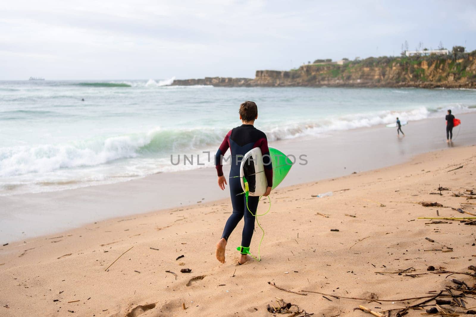 Back view of anonymous surfer in wetsuit with surfboard walking along sea waves. Active surfer in swimwear walking on sandy beach. Embodying the concept of a summer vacation.