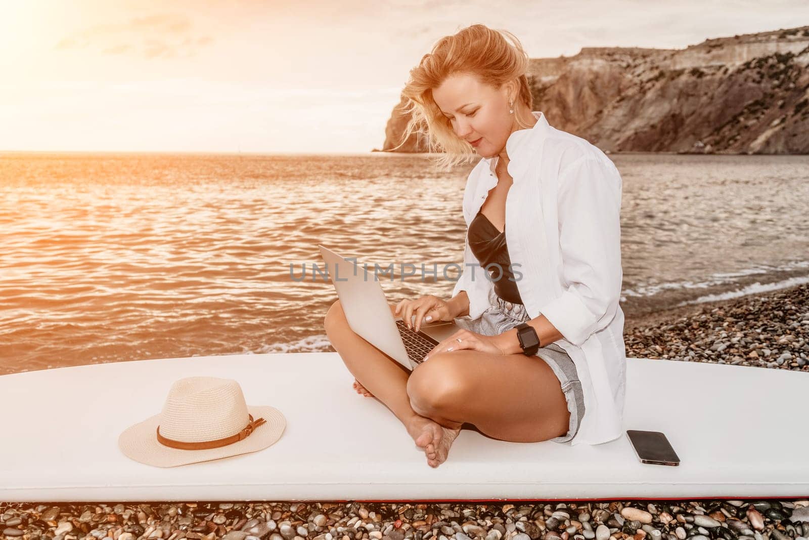 Successful business woman in yellow hat working on laptop by the sea. Pretty lady typing on computer at summer day outdoors. Freelance, travel and holidays concept.