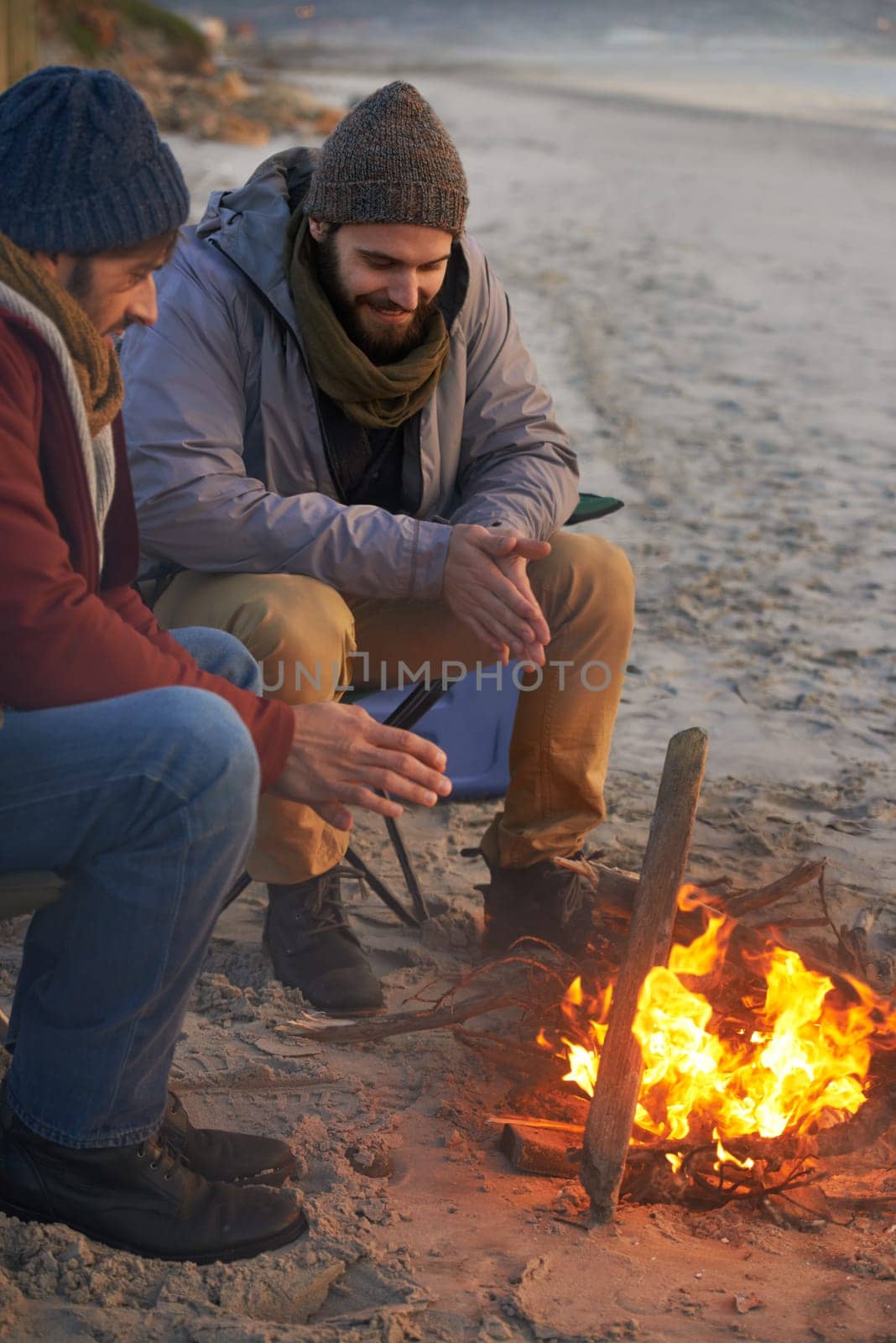 Men, sunset and campfire with cold hands, smile and happy from travel adventure and journey outdoor. Friends, ocean and sea with bonding, nature and vacation by a beach together on holiday with fire.
