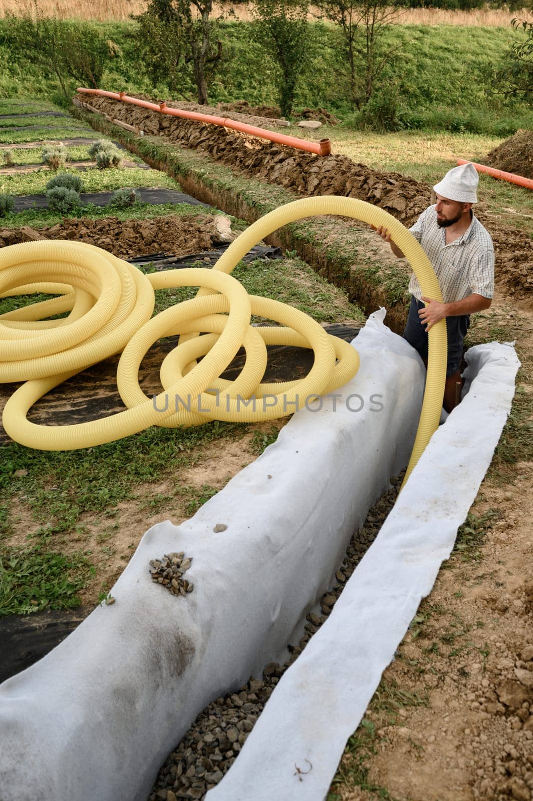 A worker carries a yellow perforated drainage pipe. Groundwater drainage works in the field by Niko_Cingaryuk