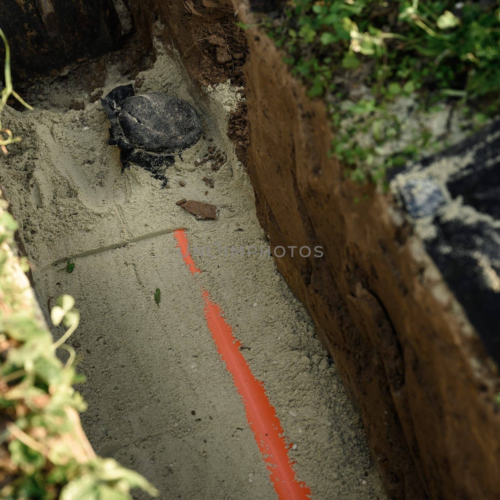 An orange reinforced sewer pipe is compacted with sand in a trench. Plumbing works on private plot. by Niko_Cingaryuk