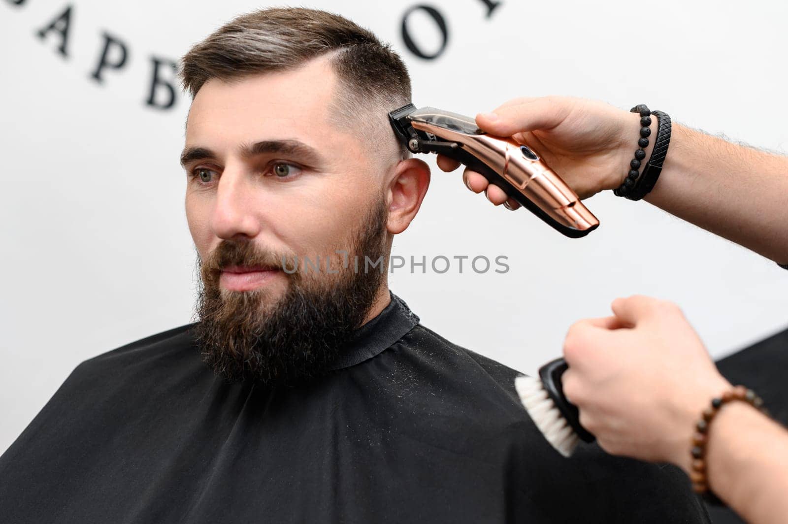 A barber cuts a man with a beard in a barber shop. Short haircut of the client with a clipper.