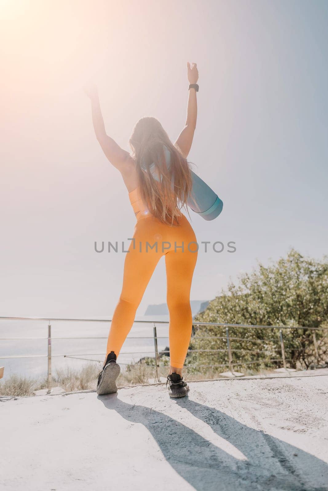 Fitness woman sea. Happy middle aged woman in orange sportswear exercises morning outdoors on yoga mat with laptop in park over ocean beach. Female fitness pilates yoga routine. Healthy lifestyle. by panophotograph