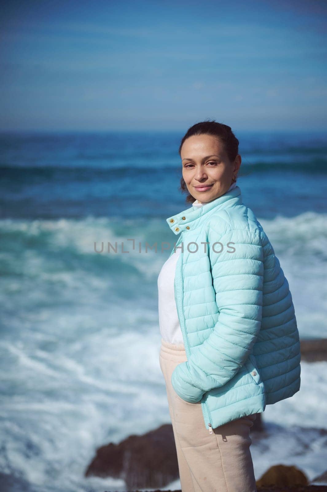 Waist up portrait of a happy smiling woman standing by ocean on the rocky cliff, looking at camera, enjoying the view of beautiful waves making white foam while crushing on the headland