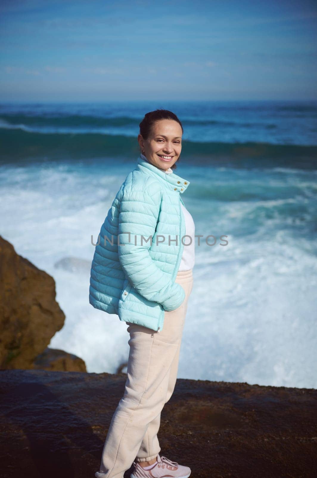 Happy smiling woman standing by ocean on the rocky cliff, looking at camera, enjoying the view of beautiful waves making white foam while crushing on the headland