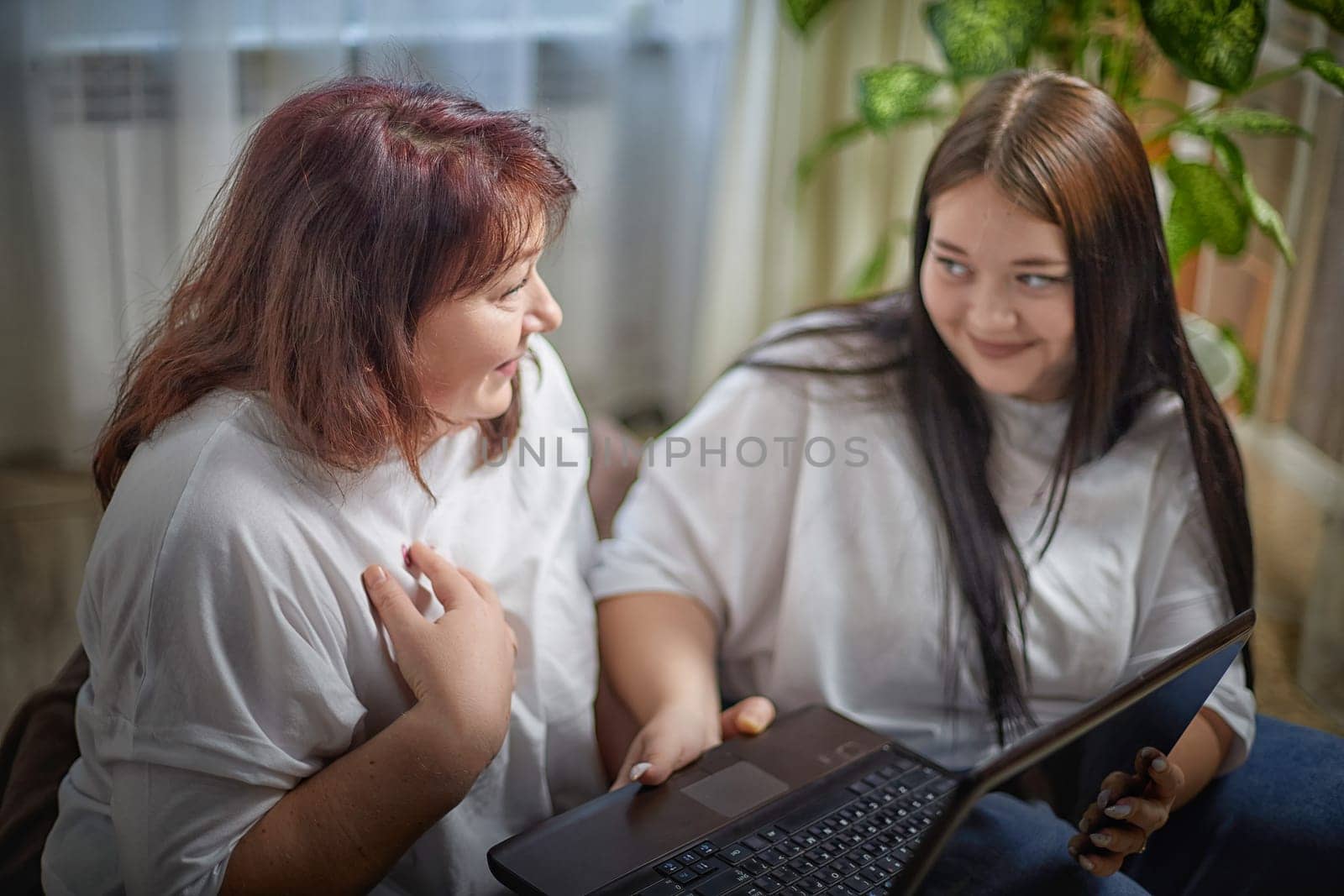 Fat funny funny adult mother and daughter with laptop indoors. A teenage girl teaches middle-aged woman modern technology. Internet, chatting by keleny