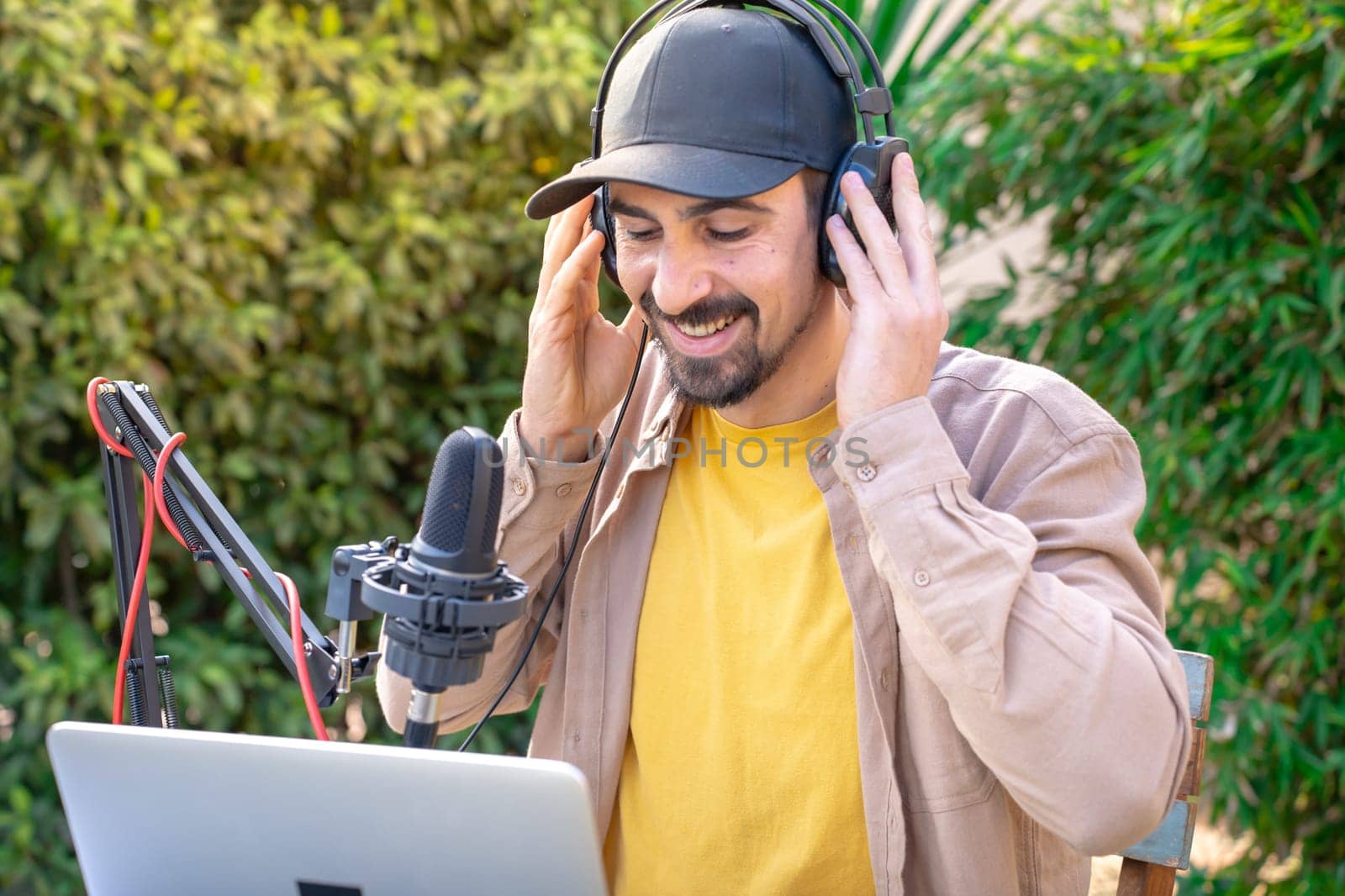 A man with a beard and moustache wearing a cap and headphones is singing and recording podcast into a microphone while using a laptop outdoor. He looks happy and relaxed, enjoying a leisurely musical event