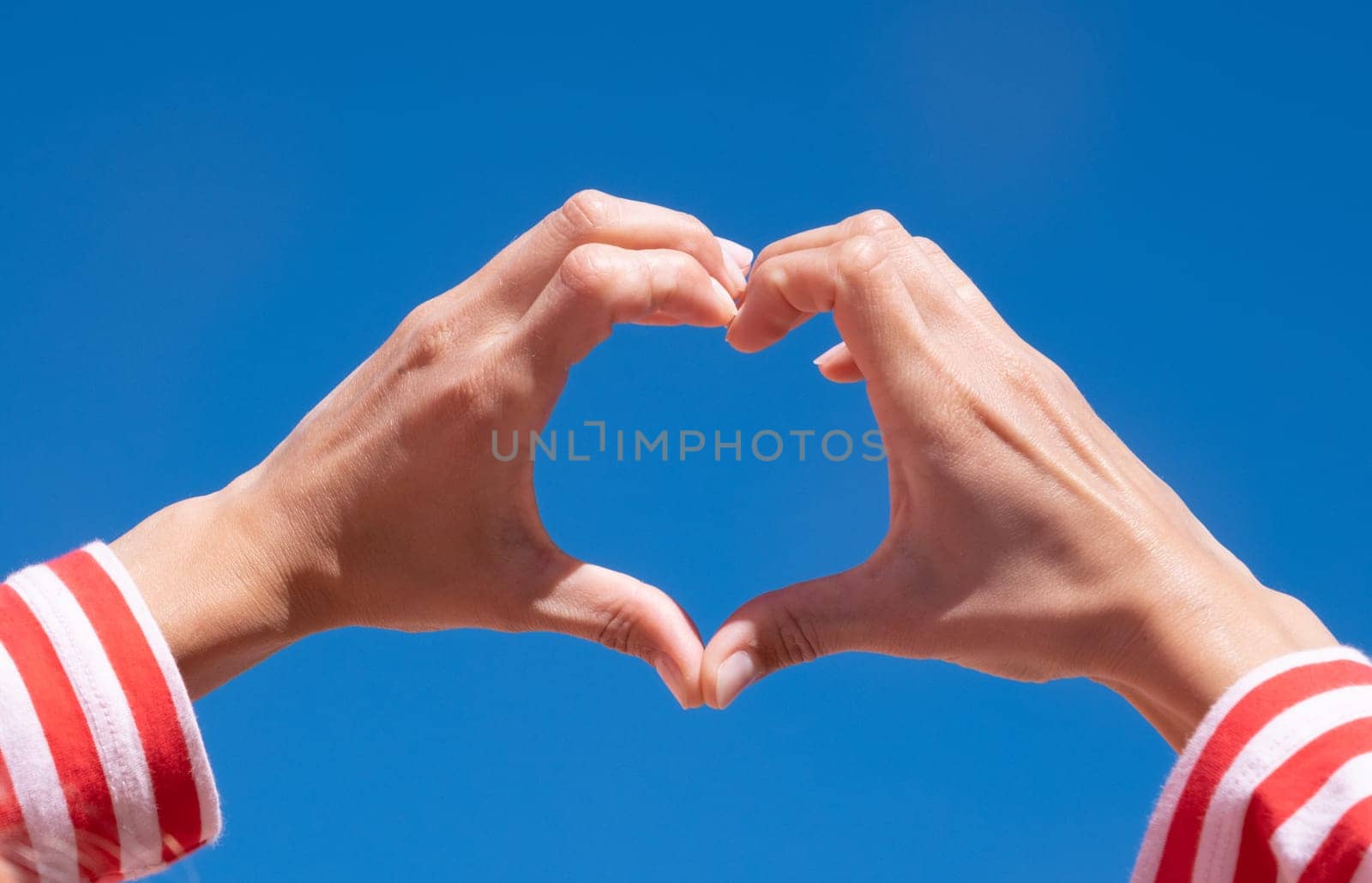 Female hands making sign Heart by fingers on sky background by Desperada