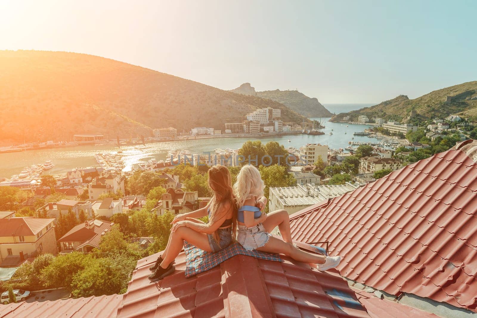 Two women sitting on a red roof, enjoying the view of the town and the sea. Rooftop vantage point. In the background, there are several boats visible on the water, adding to the picturesque scene. by Matiunina