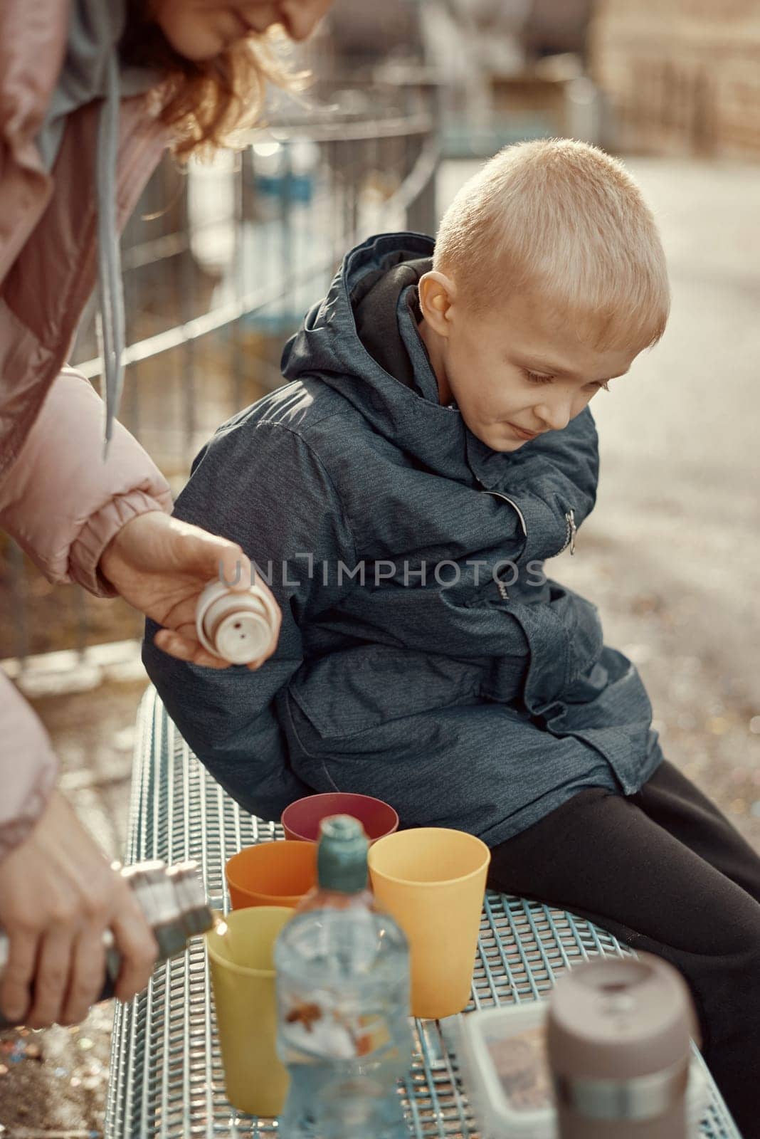 Family Picnic Delight: Cheerful 8-Year-Old Blond Boy in Blue Winter Jacket Sits on Bench While Mom Pours Tea from Thermos, Autumn or Winter. a joyful 8-year-old blond boy in a blue winter jacket, sitting on a bench while his mom pours tea from a thermos into colorful plastic cups. by Andrii_Ko