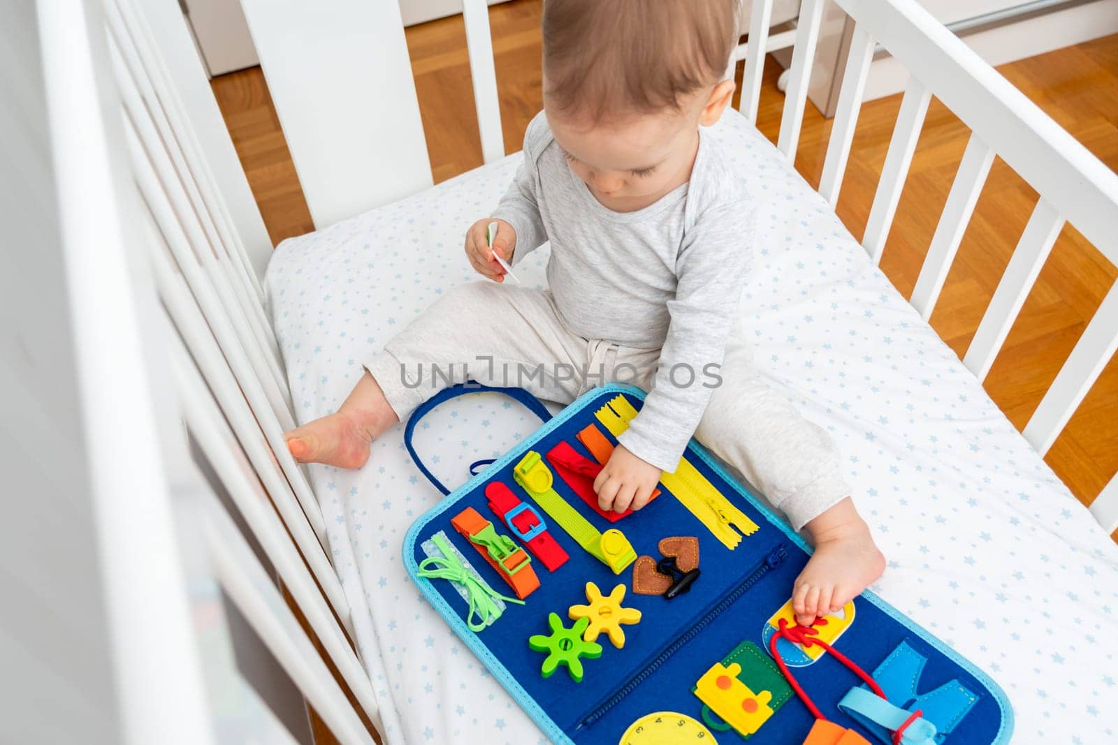 Baby girl playing with montessori busy book sitting in a crib, concept of early education by Mariakray