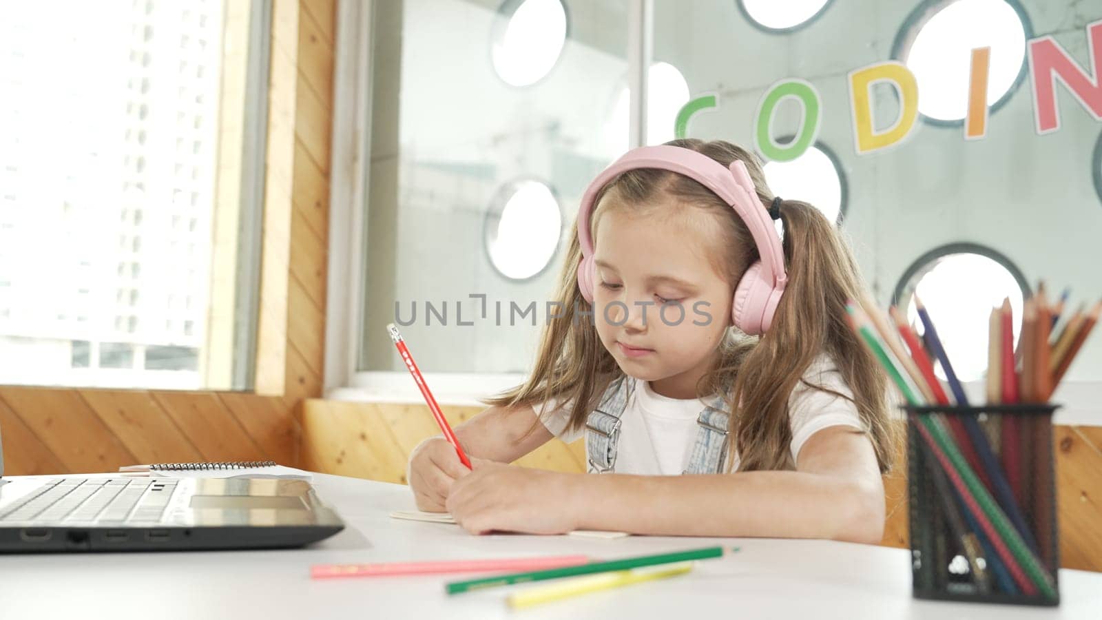 Cute smart girl doing classwork or online learning while listen to teacher. Happy student writing, drawing, working on paper while wearing headphone with laptop and colorful color pencils. Erudition.