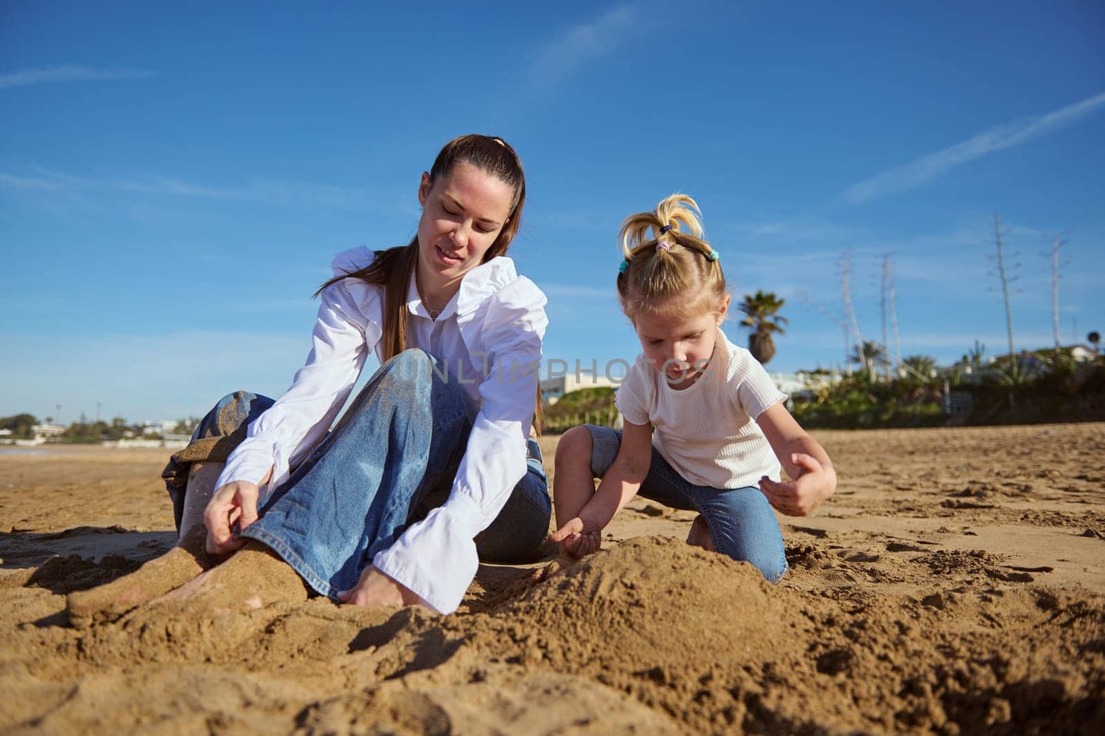 Happy mother and daughter playing together on the beach, building sandy castle