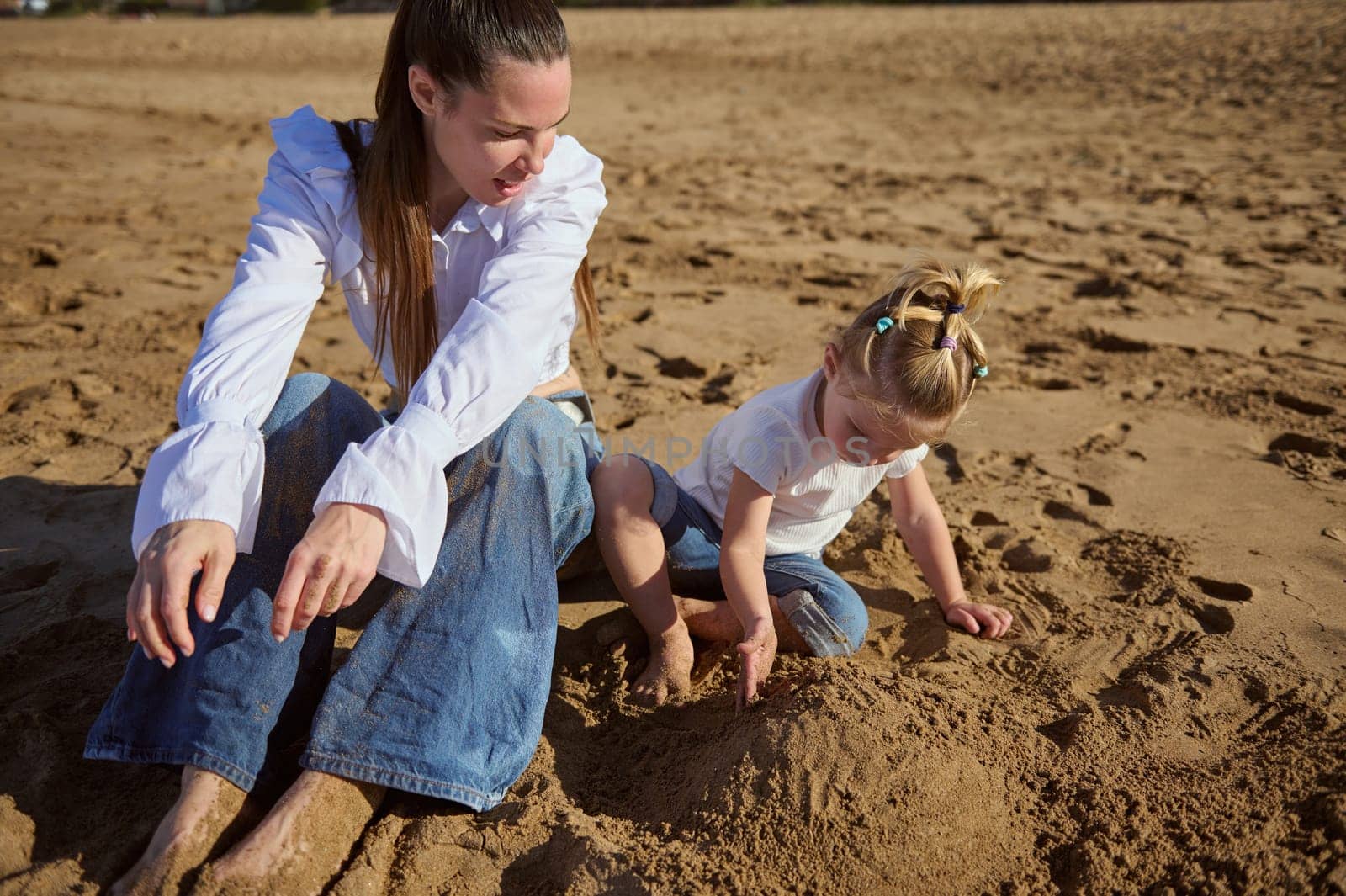 Happy family of a beautiful Caucasian single mother and her adorable little kid girl, lovely daughter sitting on the sand and playing together, building sandy castle
