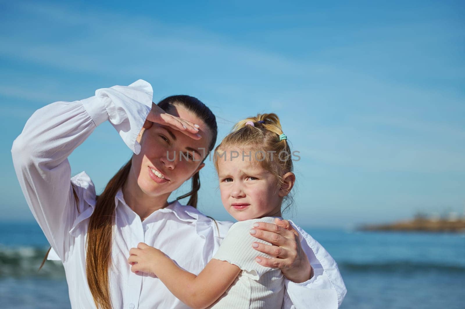 Happy caring mother holding her baby girl in her arms while walking together on the beach, looking into the distance by artgf