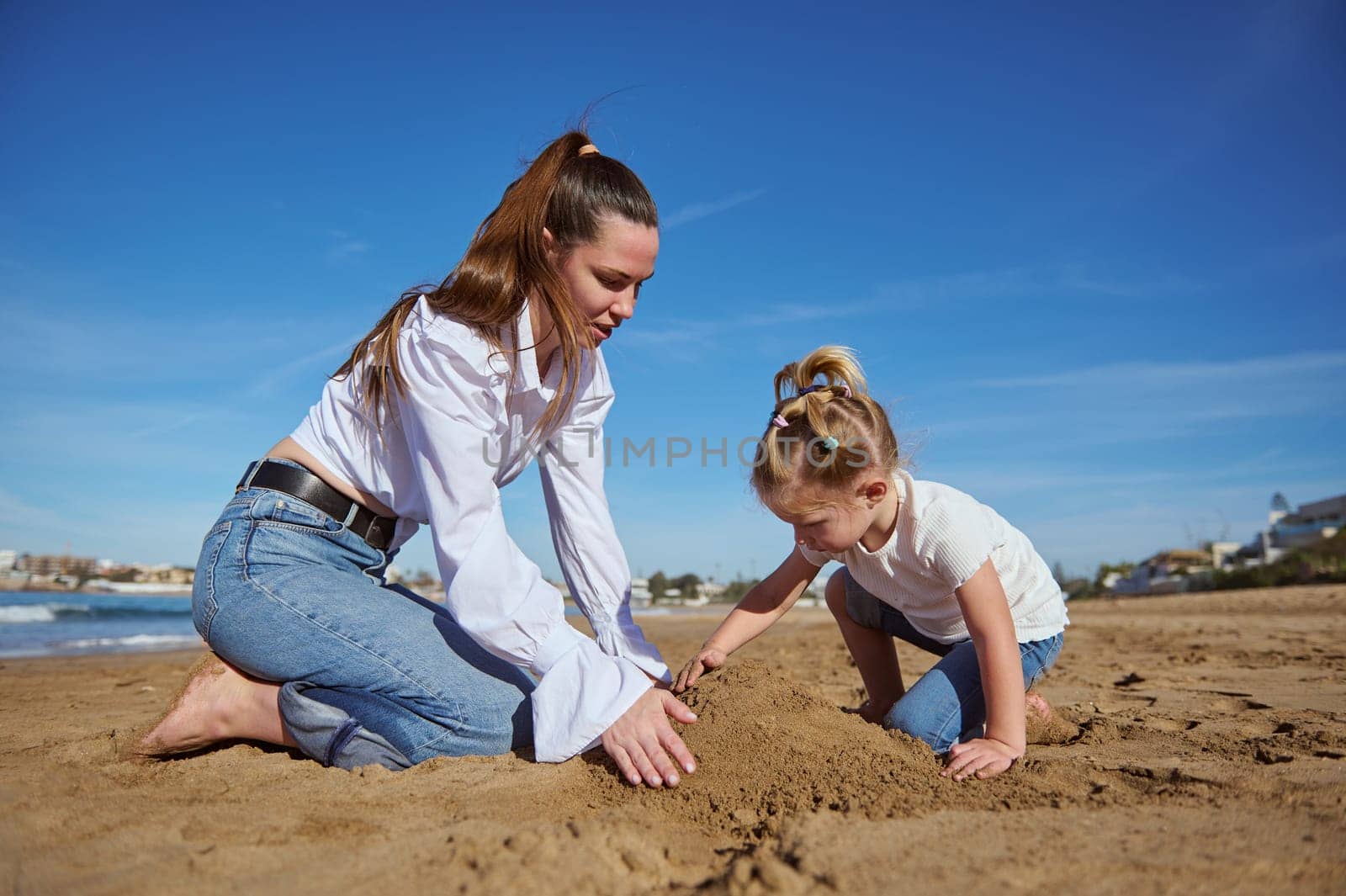 Happy family of mother and her cute little kid girl, building a sand castle on the beach on a sunny day, against the background of the sea and the sky with clouds. Mom and daughter relaxing together.