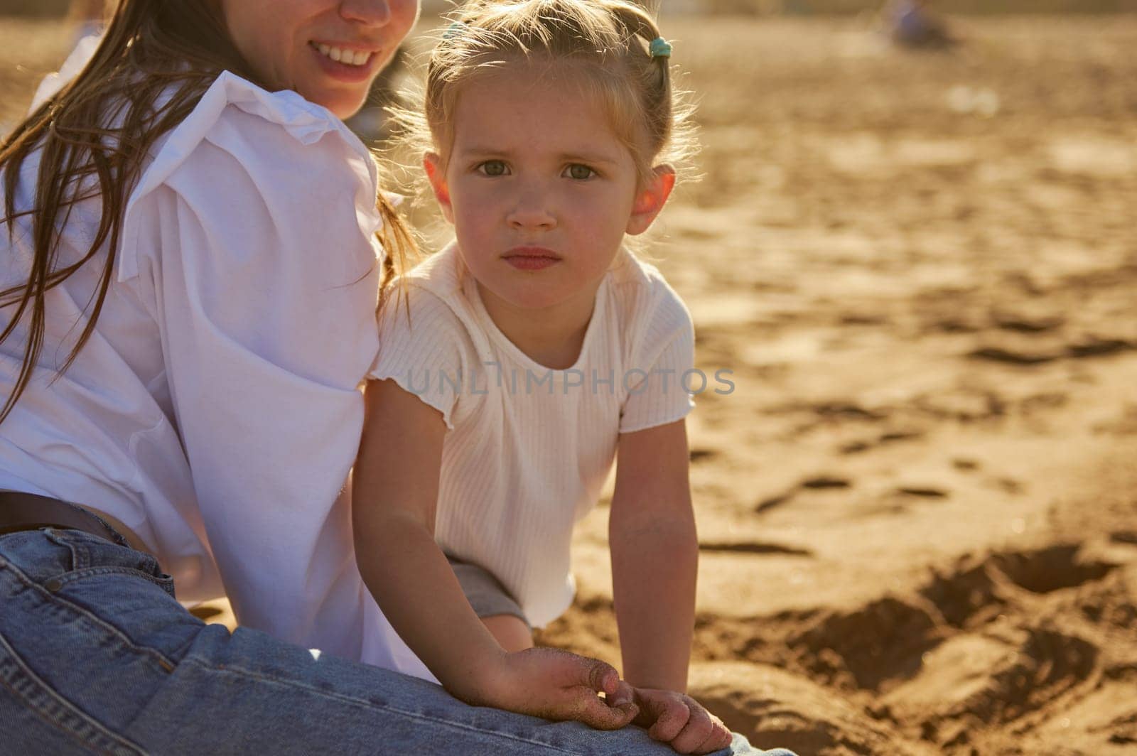 Adorable little child girl looking at camera, sitting near her loving mother on the sandy beach, playing together at sunset