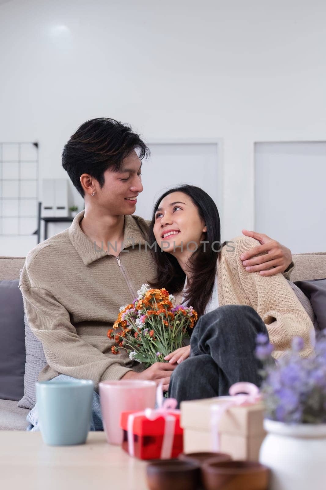 A young Asian couple gives flowers to each other on their anniversary and sits happily together in the living room. by wichayada