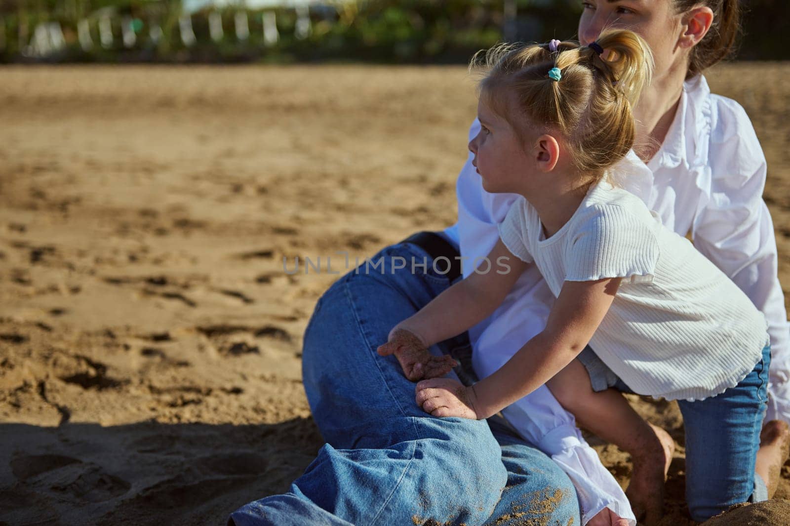Authentic portrait of family of a happy young woman, loving mother enjoying happy moments with her cute little kid girl, playing together on the sandy beach at sunset. People and lifestyle concept