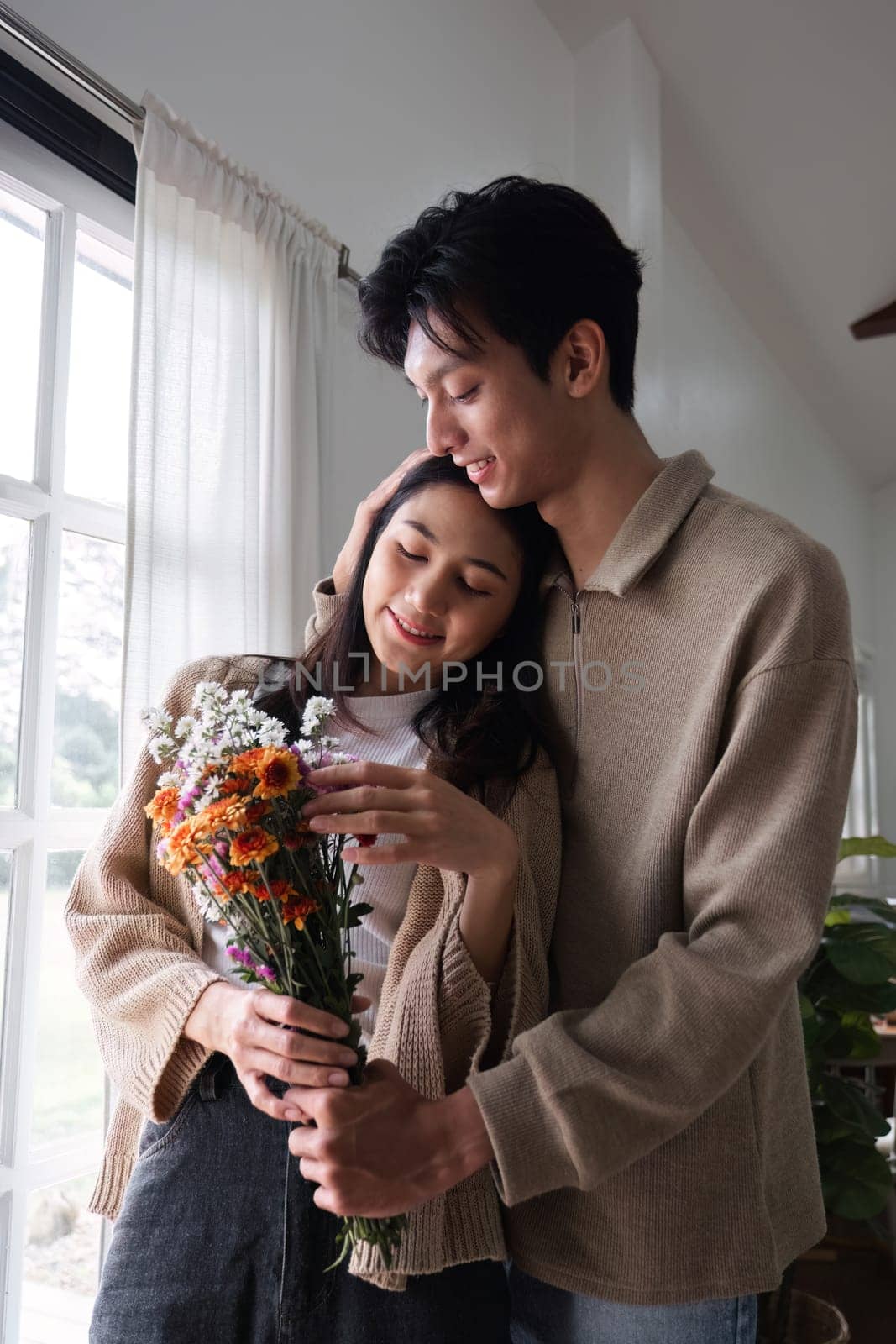 A young Asian couple gives flowers to each other on their anniversary and sits happily together in the living room. by wichayada