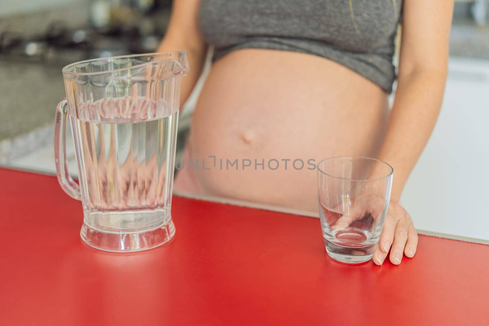 Embracing the vital benefits of water during pregnancy, a pregnant woman stands in the kitchen with a glass, highlighting hydration's crucial role in maternal well-being.