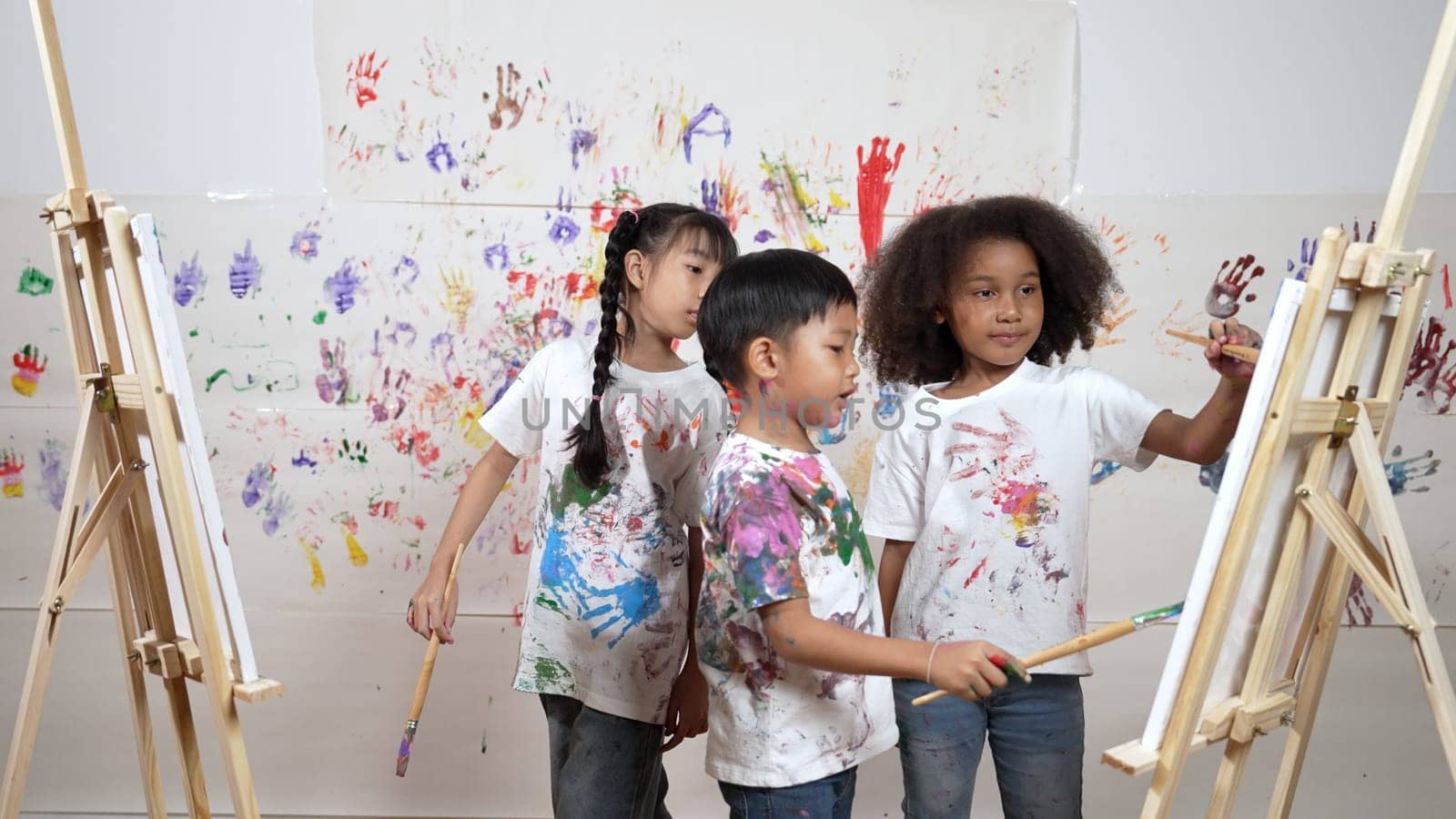 Diverse student painted or draw canvas at stained wall in art lesson. Asian girl wearing white shirt with stained color while standing at stained wall with hand print. Creative activity. Erudition.