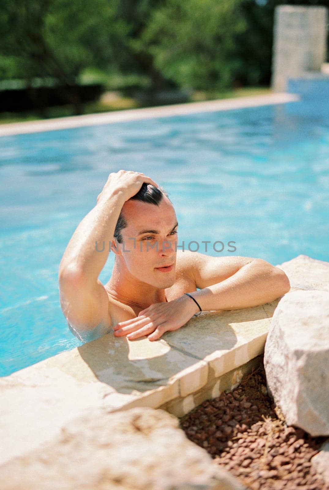 Young man runs his hand through his hair, leaning on the side of the pool and looking into the distance. High quality photo