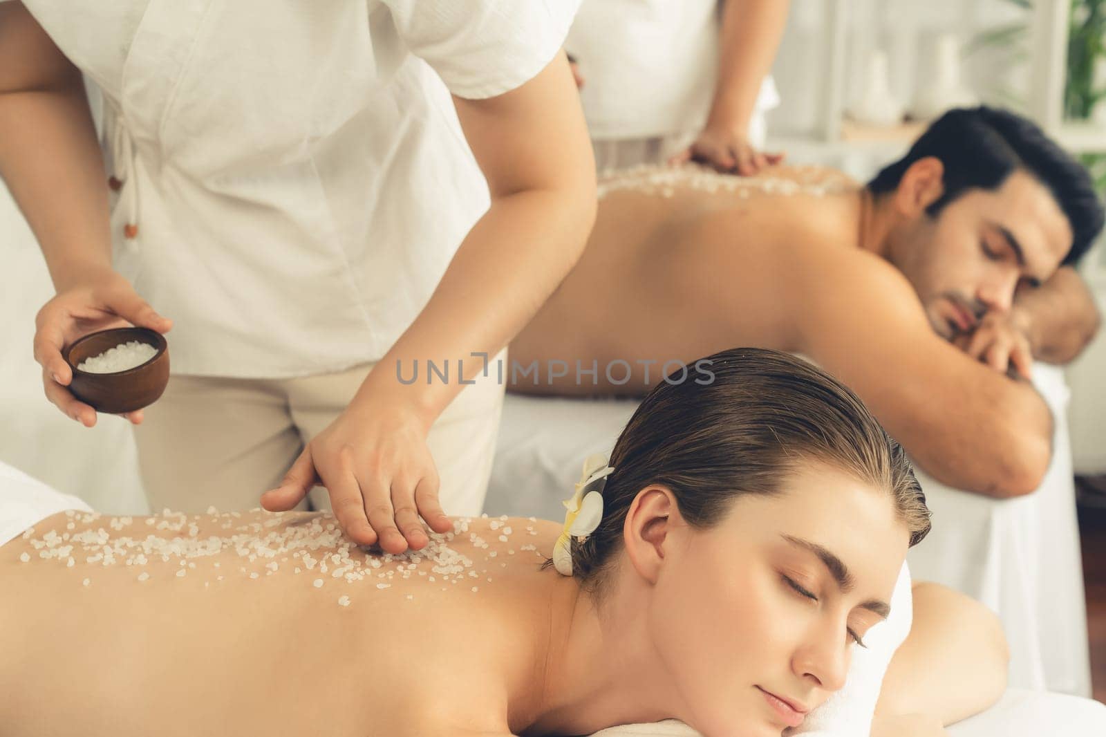 Blissful couple customer having exfoliation treatment in luxury spa salon with warmth candle light ambient. Salt scrub beauty treatment in health spa body scrub. Quiescent