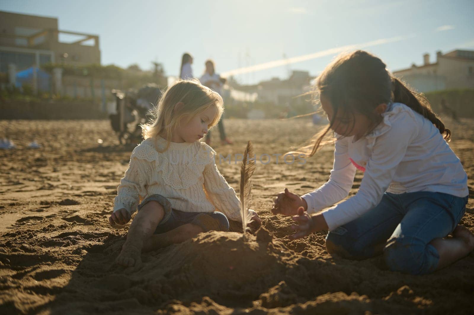 Two adorable and happy Caucasian kids girls building figures from sand while playing on the sandy beach at sunset. Happy carefree childhood. Vacations and holiday concept