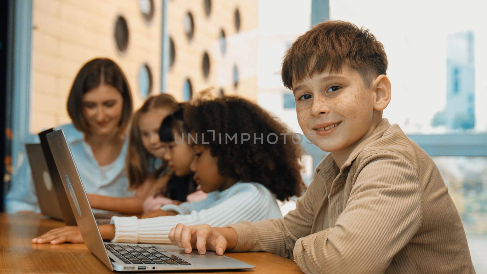 Boy looking at camera with multicultural friend learning prompt at STEM technology class. Multicultural student study about engineering code and programing system with blurring background. Erudition.