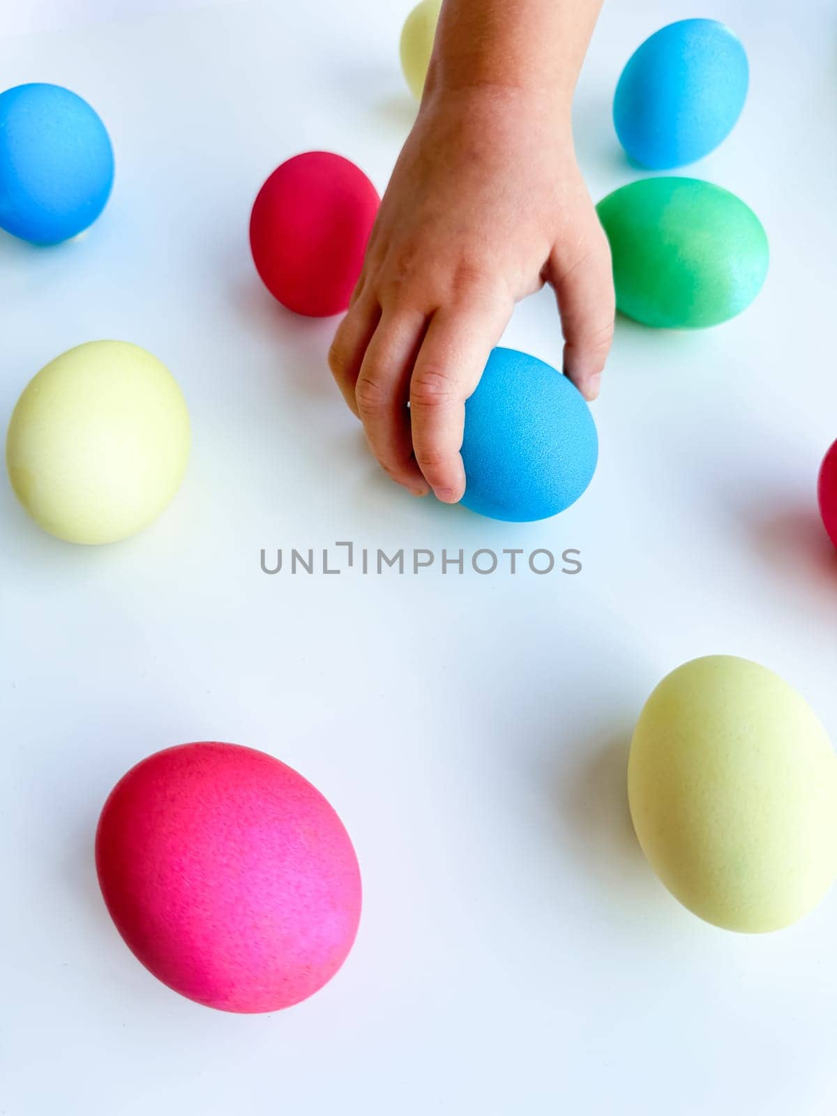 Child's hand picking up a blue Easter egg among colorful eggs on a white surface, interactive and engaging holiday activity concept. Can be used for educational content highlighting fine motor skills development in children, Easter egg hunt event promotions, family friendly activity ideas. . High quality photo