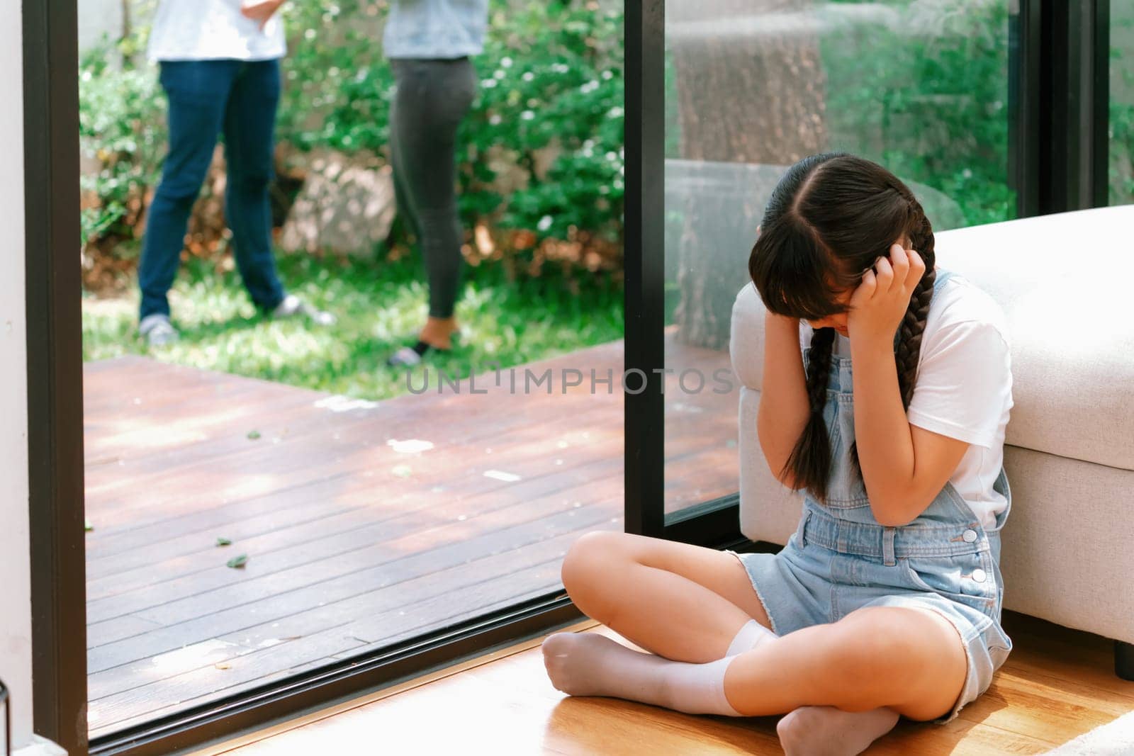 Stressed and unhappy young girl huddle in corner, cover her ears blocking sound of her parent arguing in background. Domestic violence at home and traumatic childhood develop to depression. Synchronos