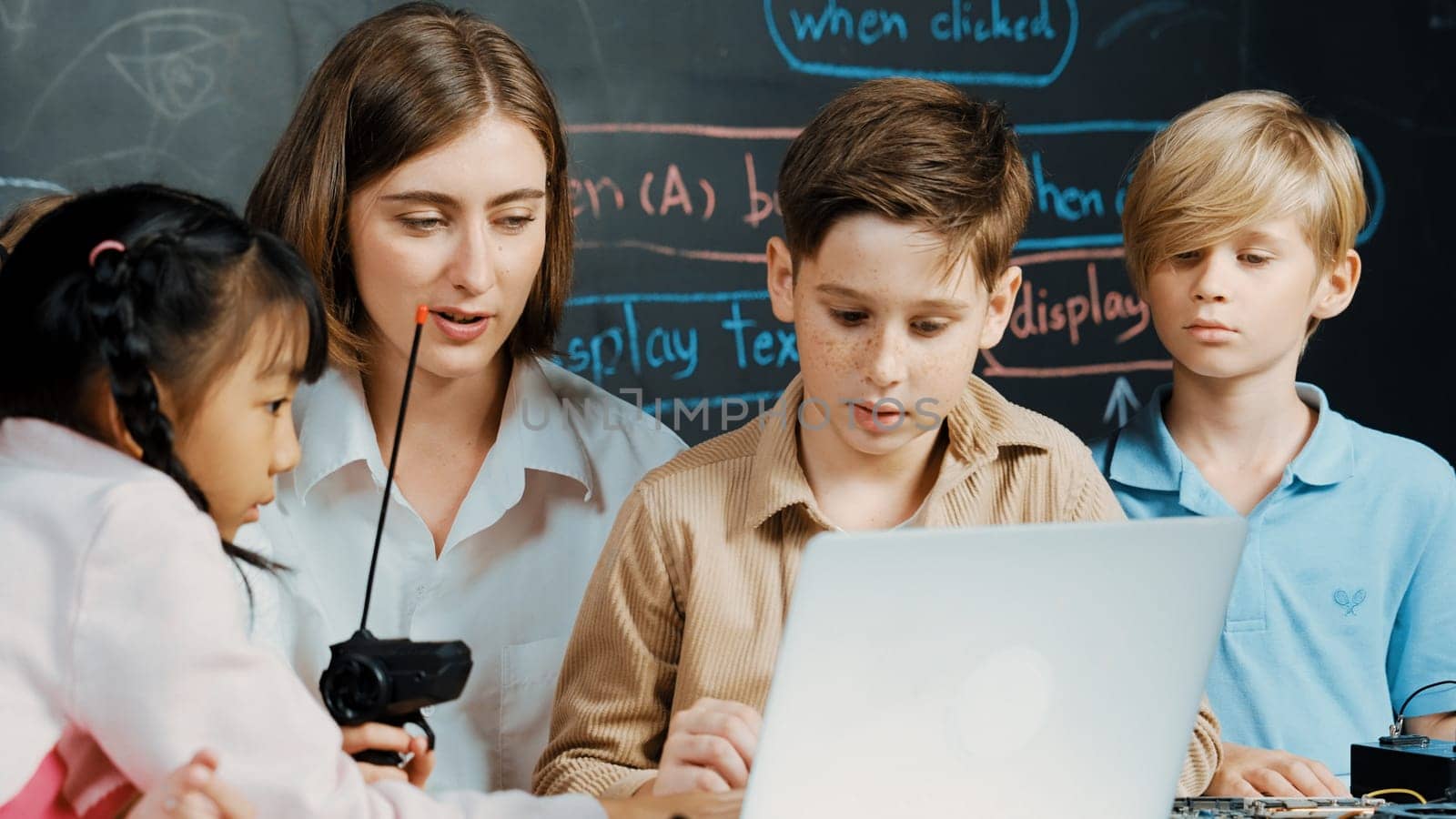 Caucasian boy using laptop programing engineering code and writing program while young caucasian teacher holding controller in STEM technology classroom at blackboard written with prompt. Erudition.