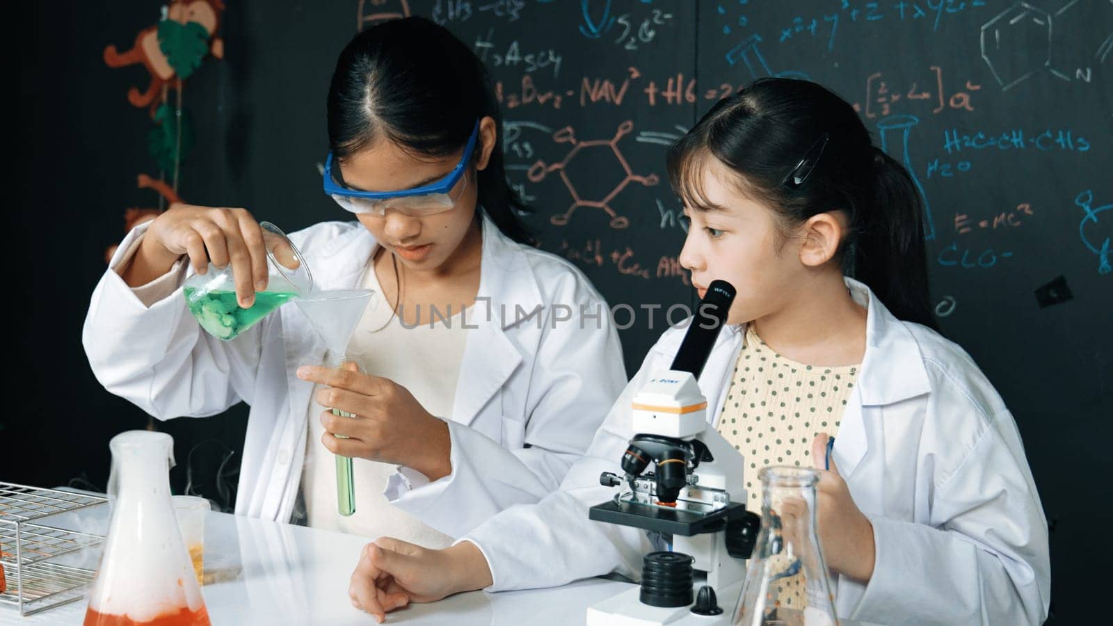 Young highschool student doing experiment by pouring sample in test tube. Girl wearing glasses and looking under microscope to inspect sample in STEM science class. Creative education. Edification.