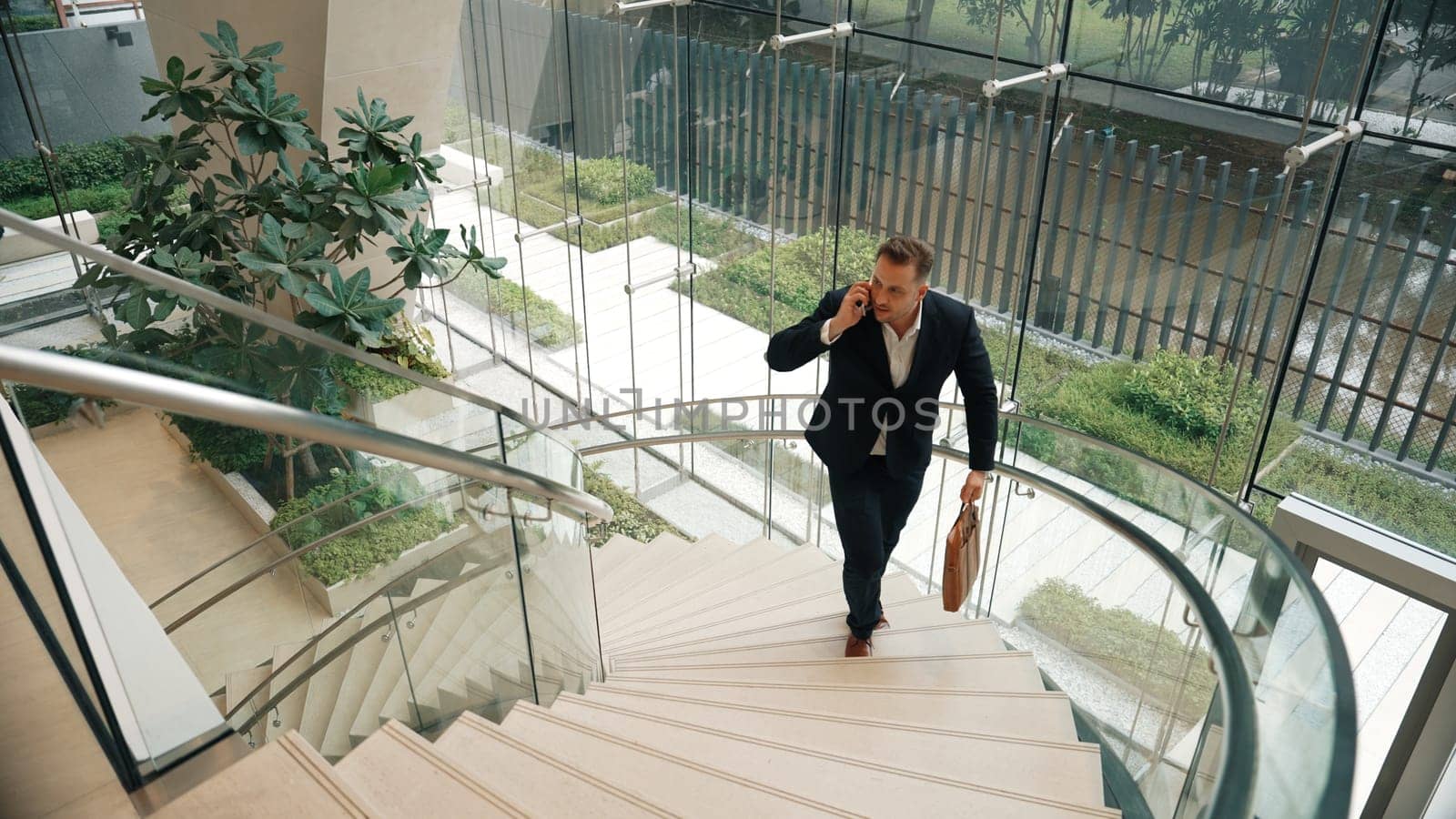 Smart businessman walking up spiral stair case and calling manager by using smart phone. Top view of manager talking marketing team by using telephone while going up stair at modern building. Urbane.