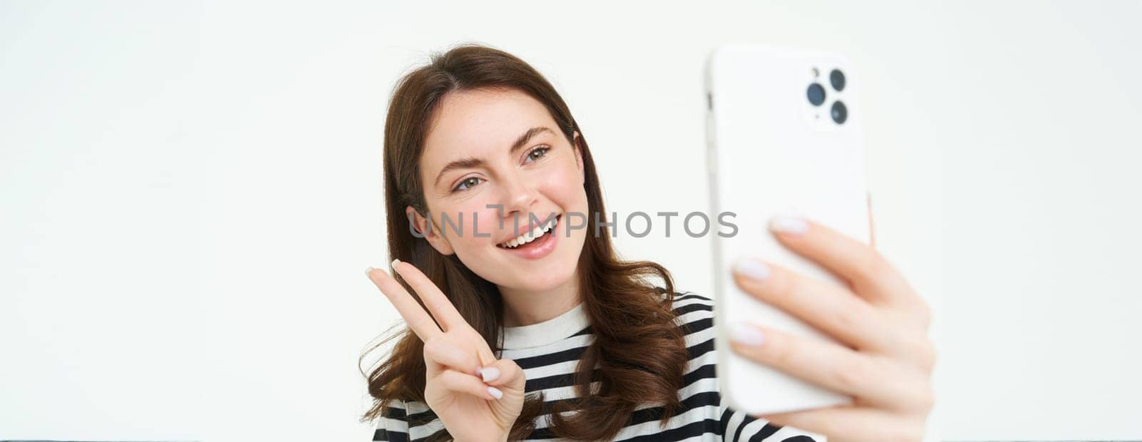 Portrait of young woman taking selfies on smartphone, posing for photo,. using mobile phone app for taking funny and cute pictures, isolated on white background.