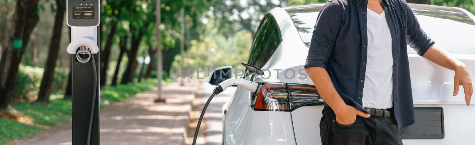 Man recharging battery for electric car during road trip travel EV car in natural forest or national park. Eco friendly travel during vacation and holiday. Panorama banner Exalt
