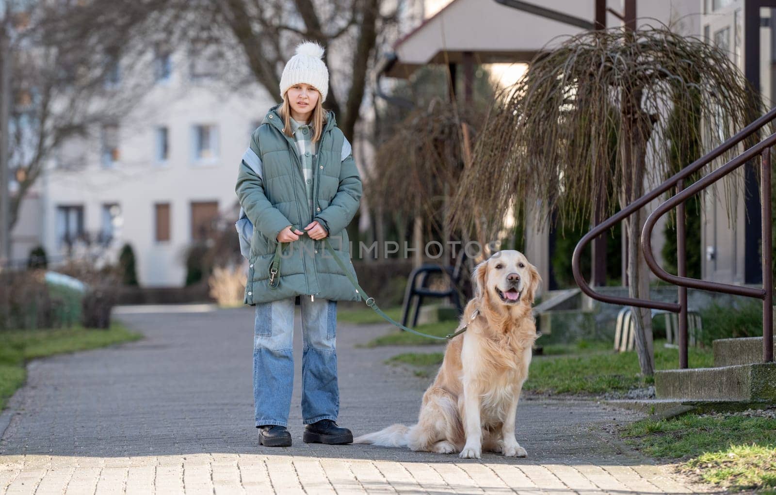 Small Girl In Green Jacket Walks With Golden Retriever On Street In Early Spring, Showcasing A Child And Dog Bond