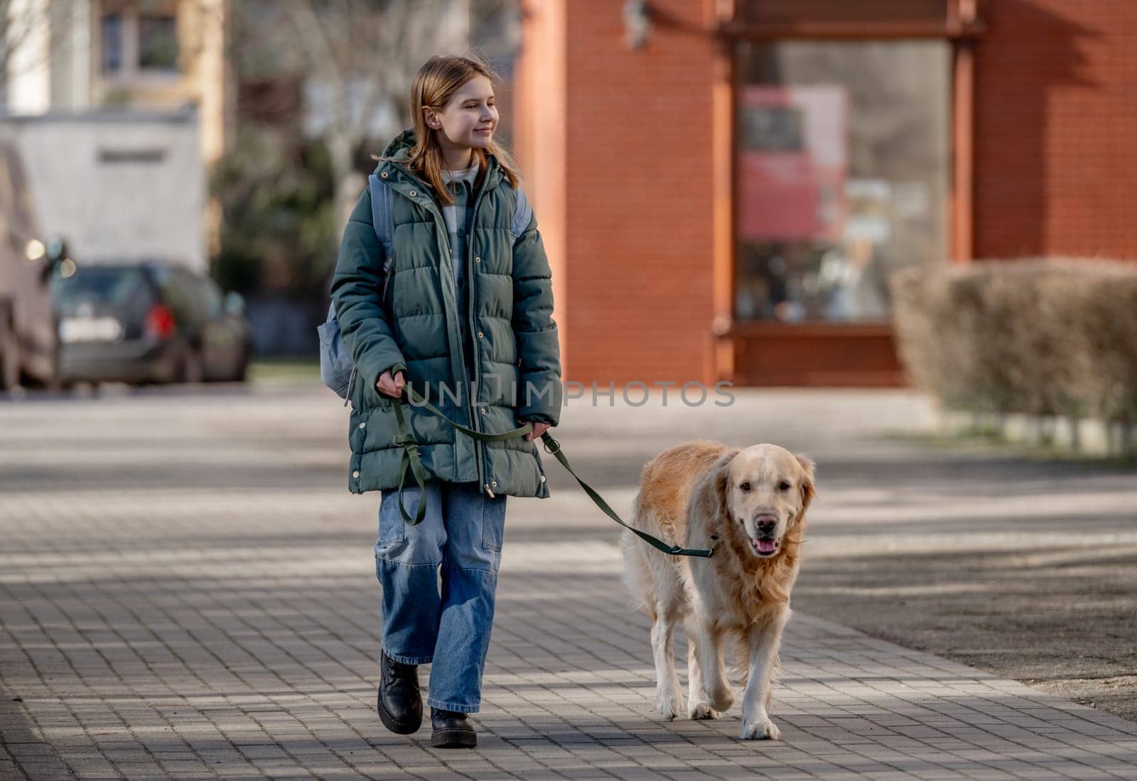 Girl In Green Jacket Walks With Golden Retriever On City Street In Early Spring