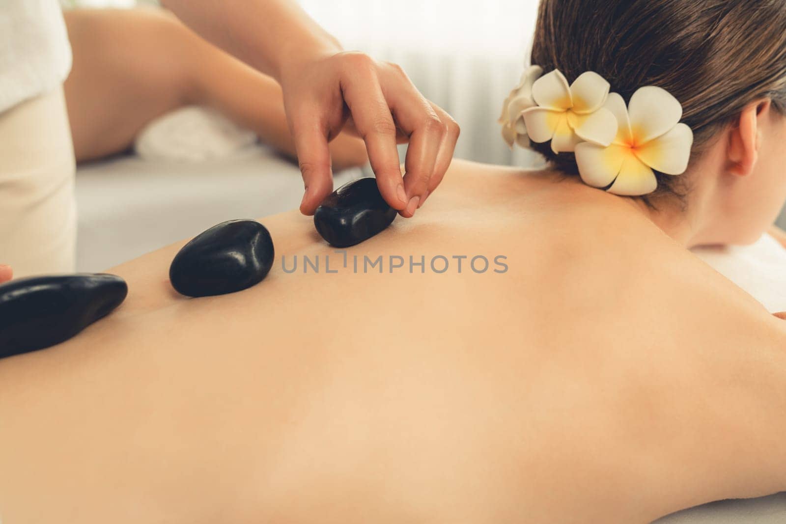 Hot stone massage at spa salon in luxury resort with day light serenity ambient, blissful couple customer enjoying spa basalt stone massage glide over body with soothing warmth. Quiescent