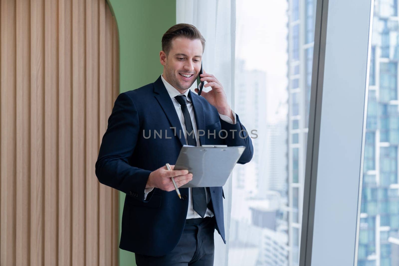 Portrait of professional businessman stand near window with skyscraper while phone calling and holding laptop. Busy executive manager holding laptop while multitasking working. Closeup. Ornamented.